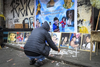 Una persona agachada enciende velas blancas sobre el suelo. En el suelo hay retratos enmarcados de hombres, mujeres y niños. Un gran cartel con fotos y texto se ve al fondo