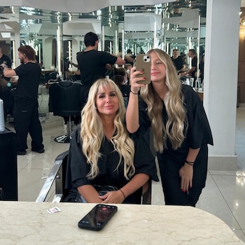 Dos mujeres rubias en batas negras se toman una selfie en un salón de belleza con espejos. Un teléfono móvil está sobre una mesa de mármol