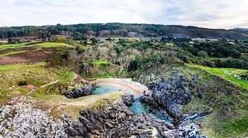 Vista aérea de una pequeña cala con playa de arena, agua turquesa, acantilados rocosos, senderos y colinas verdes con bosque al fondo.