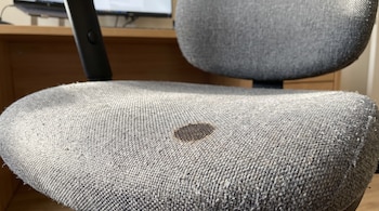 Una silla de escritorio de tela gris con una mancha circular oscura en la parte central del asiento.