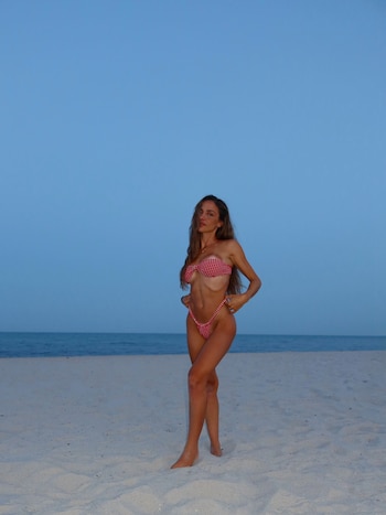 Mujer de cabello largo en bikini a cuadros rojos y blancos posa en una playa de arena clara con el mar y un cielo azul de fondo