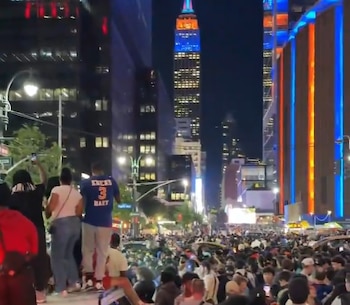 Una calle de Nueva York de noche con una gran multitud de personas. El Empire State Building y otros edificios están iluminados en azul y naranja