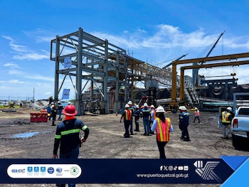 Trabajadores con cascos y chalecos reflectantes frente a una estructura metálica en construcción con grúas y maquinaria pesada