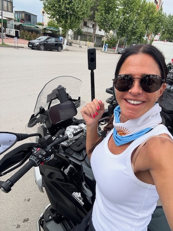 Carolina Baldini sonríe sentada en una motocicleta negra, con gafas de sol, camiseta blanca, pañuelo argentino y cámara 360 en una calle