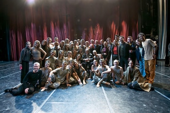 Un gran grupo de personas, incluyendo Charly García en silla de ruedas y Julio Bocca, posan en un escenario con telones rojos. Algunos llevan vestuario de ballet