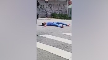 Una mujer con camiseta azul y pantalón de mezclilla yace en el asfalto frente a un paso peatonal y una persiana metálica con grafitis