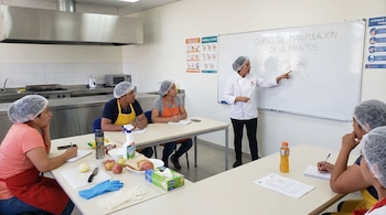Instructora en chef enseña a cinco estudiantes con gorros de red en una cocina. Señala dibujo de manos en pizarra. Mesas con frutas, pan, guantes y útiles.