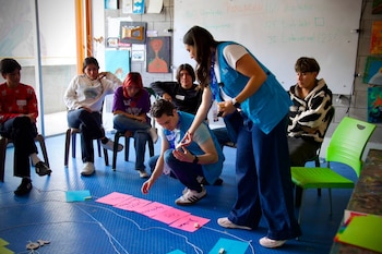 Jóvenes sentados en círculo observan a dos instructores con chalecos azules organizar tarjetas rosadas sobre el suelo de un aula