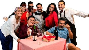 Un grupo de personas posa sonriendo alrededor de una mesa con mantel a cuadros, botellas de vino, una panera y copas