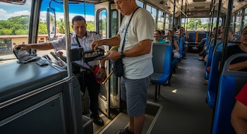 Un hombre paga con monedas a un conductor de autobús en el interior de un autobús lleno de pasajeros, con paisajes verdes visibles por la ventana.