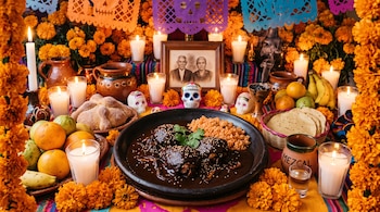 Plato con mole y arroz rodeado de flores de cempasúchil, velas encendidas, pan de muerto, frutas, calaveritas de azúcar y un retrato familiar.