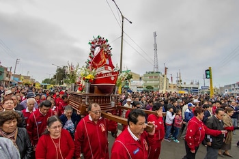 Gran multitud de personas acompaña una procesión llevando en andas un bote rojo decorado con flores y una figura religiosa por una calle urbana