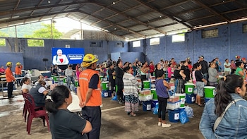 Interior de un almacén con un grupo de personas. Hay cubos y cajas de ayuda apilados, y una pantalla mostrando a Maryflor Morates al fondo