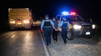 Dos policías hondureños escoltan a una mujer esposada hacia una patrulla con luces azules y rojas en carretera rural oscura, con un camión amarillo estacionado.