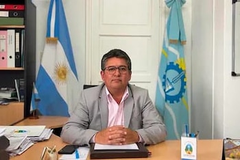 Hombre con anteojos y saco gris sentado ante un escritorio con las manos juntas, entre la bandera de Argentina y una bandera institucional