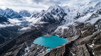 Vista aérea de un lago turquesa rodeado de morrenas oscuras, rocas, hielo y montañas nevadas con nubes.