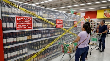 Pasillo de supermercado con estanterías de licores cubiertas con plástico y cinta amarilla de "CAUTION", y letreros de "LEY SECA". Compradores con carritos al frente.