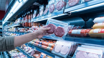 Una mano sostiene un envase de plástico con carne frente a estanterías refrigeradas llenas de embutidos y carnes envasadas.