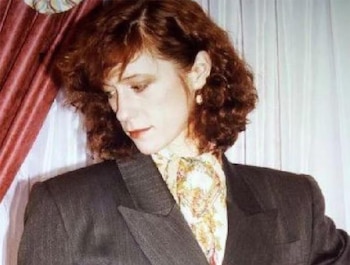Retrato de Shelly Miscavige, mujer de piel clara con cabello rizado castaño rojizo, vistiendo un traje oscuro y una blusa estampada, mirando hacia abajo. Fondo con cortinas rojas y blancas