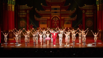 Veinticinco artistas vestidos con trajes de colores, incluyendo un hombre de rojo al centro, en un escenario con un fondo que simula un templo chino