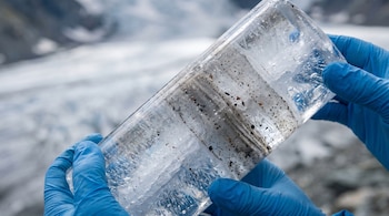 Manos con guantes azules sostienen un cilindro de hielo transparente con capas y partículas oscuras, sobre un fondo de glaciar borroso.