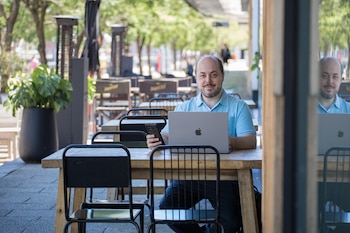 Un hombre barbudo sonríe a la cámara sentado en una mesa de madera con un portátil y un teléfono móvil; sillas metálicas y otras mesas se ven al fondo