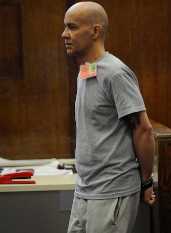 Un hombre calvo con camiseta y pantalón gris, esposado y con una credencial, mira hacia la izquierda en una sala de tribunal