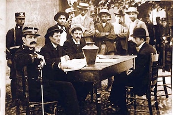 Foto sépia de homens idosos, alguns uniformizados, reunidos em torno de uma mesa de madeira com uma ânfora de metal, em uma seção eleitoral