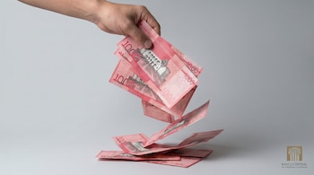 Una mano genérica suelta billetes rosados de 1,000 pesos dominicanos sobre una superficie gris claro. El logo del Banco Central es visible.