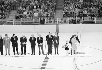 Fotografía en blanco y negro con hombres de traje y una mujer cortando una cinta en una pista de hielo ante el público