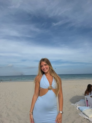 Joven rubia sonríe con vestido azul claro en la playa. El mar y el cielo nublado están al fondo, con una mesa y cojines en la arena a la derecha