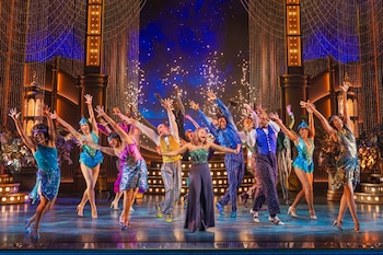 Un grupo numeroso de artistas en un escenario con vestuario colorido y brillante, decorado con elementos arquitectónicos y luces, junto a efectos de chispas