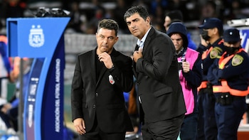 Marcelo Gallardo y Matías Biscay vestidos de traje oscuro al borde de una cancha de fútbol junto al banco de suplentes