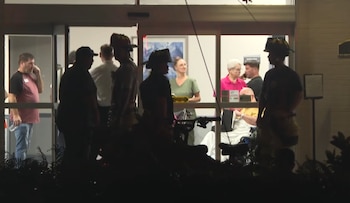 Personas en el interior de un edificio. Tres bomberos con casco, civiles, algunos ancianos y uno en silla de ruedas, por la noche