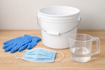 Una cubeta blanca, guantes azules desechables, un cubrebocas azul y una taza medidora transparente con agua sobre una mesa de madera clara.