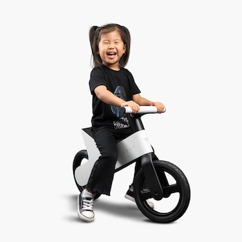 Niña sentada sobre una bicicleta de equilibrio Tesla blanca y negra con manubrios blancos, vistiendo camiseta negra, pantalón negro y zapatillas negras