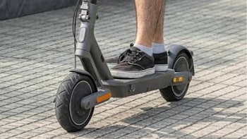 Pies y piernas de una persona sobre la plataforma de un patinete eléctrico gris que circula sobre adoquines