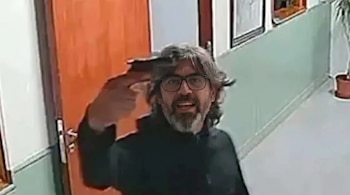 Un hombre con cabello gris, barba y gafas apunta una pistola negra a su cabeza, con la boca abierta, en un pasillo con puerta naranja y paredes claras.