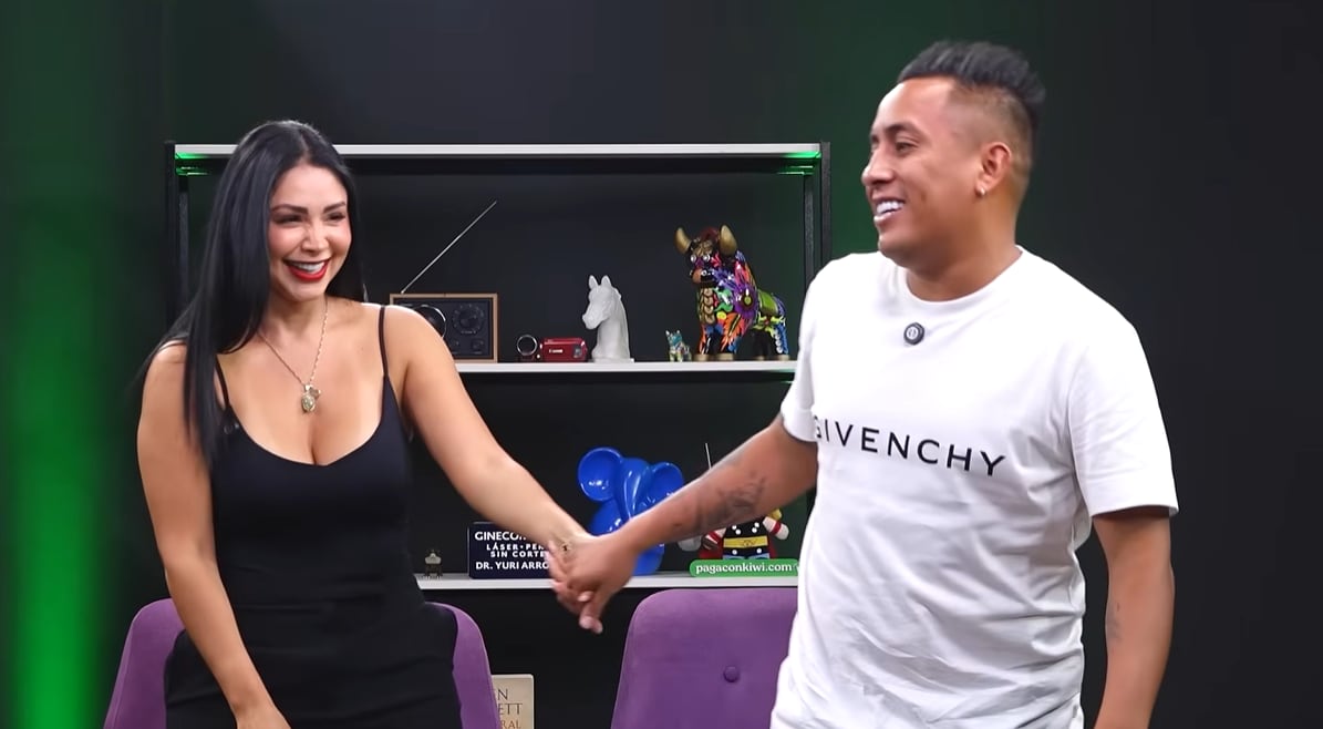 Christian Cueva y Pamela Franco aparecen sonrientes y tomados de la mano en un set de televisión con estantes decorados. (YouTube /Tampoco Tampodcast)