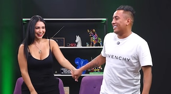 Hombre y mujer sonríen, tomados de la mano. Fondo negro y verde con estantes que sostienen adornos, una radio y partes de sillas púrpuras