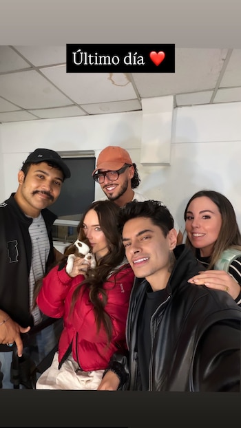 Cinco personas sonrientes, tres hombres y dos mujeres, posan en un interior. Una mujer en chaqueta roja sostiene un cachorro pequeño de pelaje blanco y marrón