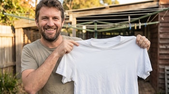 Un hombre barbudo sonríe mientras sostiene una playera blanca limpia delante de él, con un tendedero y una valla de madera al fondo.