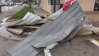 Metal corrugado gris en acera y calle. Cartel de 'STOP' rojo y tablón entre restos. Vehículos y personas al fondo