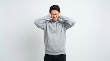 Un hombre asiático de pie, con sudadera gris y pantalón oscuro, se cubre los oídos con las manos, los ojos cerrados y el rostro contraído sobre un fondo blanco.