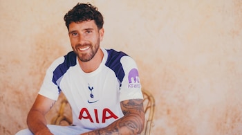Hombre joven sonriente con cabello rizado y barba, vistiendo camiseta blanca y azul marino del Tottenham Hotspur con el logo de Kraken, mostrando tatuajes en los brazos.
