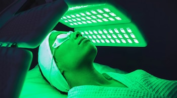 Persona acostada con protectores oculares y una toalla en la cabeza, recibiendo luz verde de dos paneles luminosos sobre su rostro.