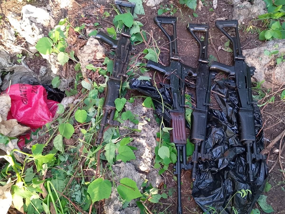 Cuatro fusiles de asalto permanecen sobre la tierra y la vegetación, junto a bolsas plásticas negras y una roja en un área exterior. (Ejército de Guatemala)