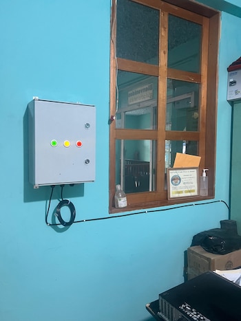 Panel de control gris con luces verde, amarilla y roja en pared azul. Ventana de madera, cableado, botella de gel, certificado y objetos en el suelo