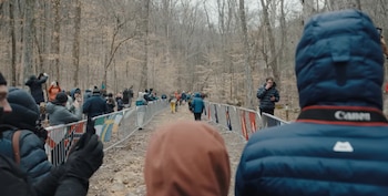 Personas con abrigos e instrumentos de fotografía permanecen tras vallas con banderas a lo largo de un sendero en un bosque de árboles secos