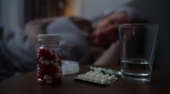 Un frasco con cápsulas rojas y blancas, blísteres de pastillas y un vaso de agua sobre una mesa de noche, con una persona al fondo